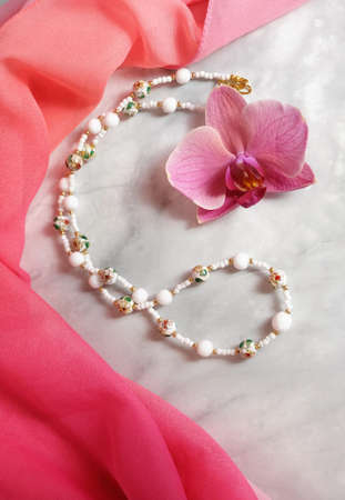 Necklace made of glass beads and couazonne beads on a background of pink chiffon drapery and orchid flower, composition with women's jewelry, selective focus, vertical orientation.の写真素材