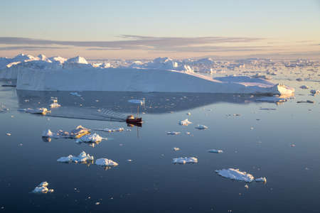 Ilulissat, Greenland - July 7, 2018: A red fishing boat sailing in between icebergs on Ilulissat Icefjord.のeditorial素材