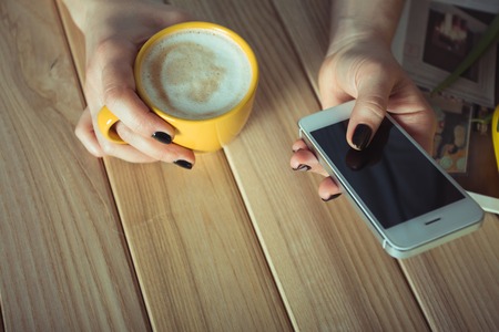 Yellow cup in the hands of women. Gel nail manicures. Phone and the magazine on the tableの写真素材
