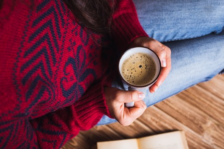 young woman in a red sweater holding a cup of coffee and reading a bookの写真素材