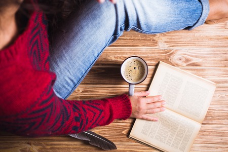 young woman in a red sweater holding a cup of coffee and reading a bookの写真素材