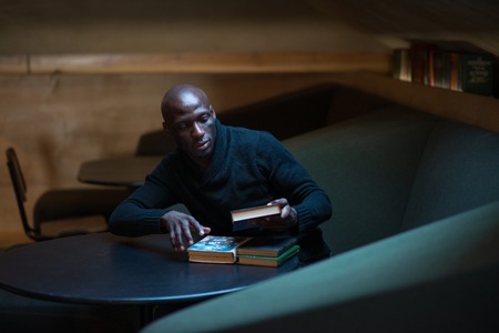 A young Arican man is reading a book in a room at a table. student, businessman