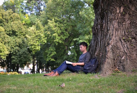 young male guy of asian appearance student sitting under tree with a book and laptop, learn college university backpackの写真素材