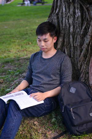 young male guy of asian appearance student sitting under tree with a book and laptop, learn college...