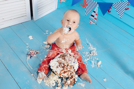decoration for the boys birthday, smash the cake in a nautical marine style. stylized birthday ship photo shoot. Cheerful boy eats and break a cake with his hands on the first holidayの写真素材