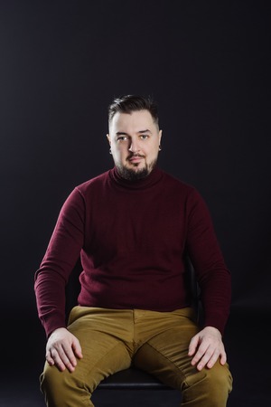 man with a beard in casual clothes poses on an isolated dark won, in a burgundy sweater in the...