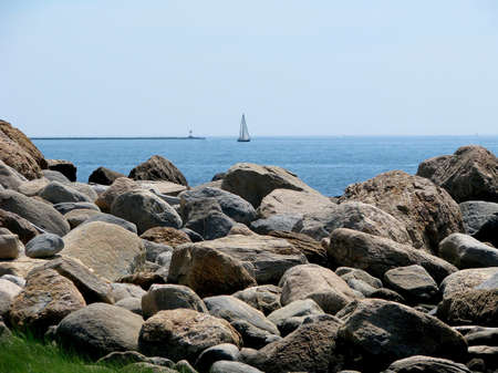 Rocky shoreline with sailboat in the background. Coast of Hammonasset Beach State Park in Madison....