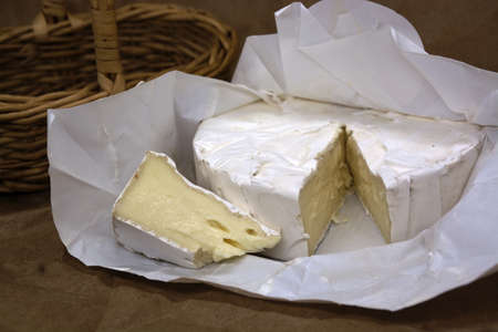Fresh brie cheese and one slice on white paper. Camembert cheese with wooden basket on background.の写真素材