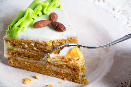 Slice of homemade carrot cake with cream cheese frosting on white plate. Person cuts cake with spoon