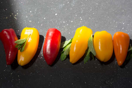colorful mini peppers and mint leaves on black sunny background. Above view of yellow, red and orange peppersの写真素材