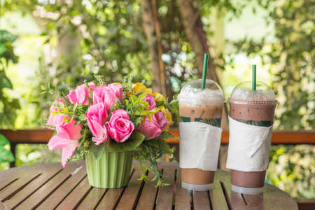 Beautiful artificial rose bouquet in vase with ice coffee and chocolate on wooden tableの写真素材
