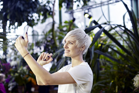 Beautuful woman with blonde short hair taking photo by phone in green park or botanical garden