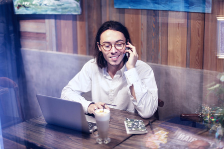 Attractive man in white shirt talking by phone and working on laptop in cafe or restaurant, having...