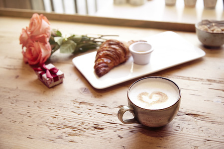 Romantic breakfast, Valentine's Day celebrating. Present box, rose flowers, fresh croissant, coffee on wooden table. Focus on cup. Daylight from window.の写真素材