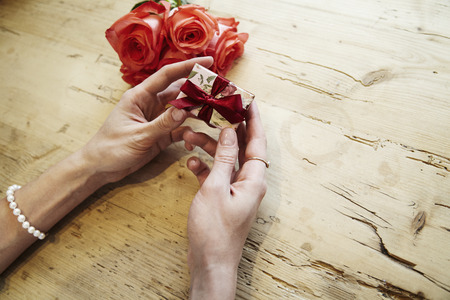 Small present box with bow in beautiful woman hands. Focus on bow. Red roses flowers behind on...