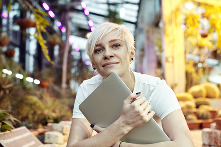 Beautiful hipster woman sitting in park or botanical garden, pensive look, smiling, thinking about things. Holding closet laptop.の写真素材