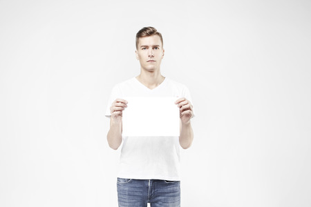 Handsome guy standing with blank sheet in hand, isolated on white background, wearing jeans and t-shirt. Free available space for layout textの写真素材