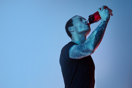 Fashion portrait of sport fit attractive man drinking water from bottle, tattooed hands, hipster look. Color flash studio lightの写真素材