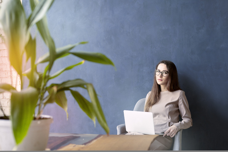 Beautiful businesswoman sitting in loft office using laptop on knees. Look and smile. Dark blue wall background, day lightの写真素材