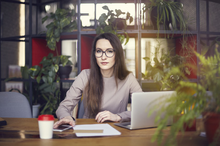 Beautiful girl wearing eye glasses in coworking studio. Using laptop and smartphone at the wooden table. Concept of young people working with mobile gadgets. Modern office blurred on background. Horizontal Beautiful girl wearing eye glasses in coworking studio. Using laptop and smartphone at the wooden table. Concept of young people working with mobile gadgets. Modern office blurred on background. Horizontal
