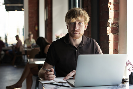 Handsome ginger caucasian man writing in notebook using laptop while sitting in cafe having coffee. Concept of young business people working in public space or co-working.の写真素材