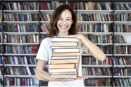 Woman model college student with books at library holds bunch of books, looks smart, smiling to camera. bookshelves at the library. Knowledge and self-developmentの写真素材