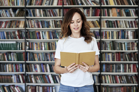 Portrait of smiling woman model reading text with opened book in a library, bookshelf behind, long hair. Hipster college student lady.の写真素材