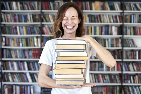 Woman model college student with books at library holds bunch of books, looks smart, smiling to camera. bookshelves at the library. Knowledge and self-developmentの写真素材