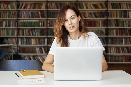 Young woman with long hair working on laptop at co-working office or library, bookshelf behind. Businesswoman reading a book, looks thoughtfullyの写真素材