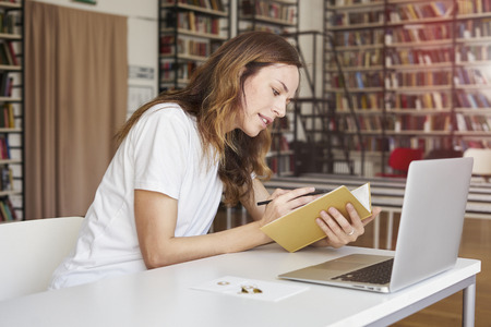 Young woman working on laptop at co-working office or library, bookshelf behind. Businesswoman writing, looks smartの写真素材