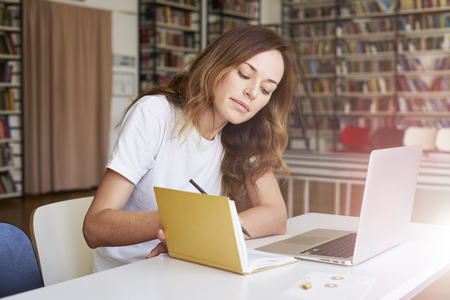 Young woman working on laptop at co-working office or library, bookshelf behind. Businesswoman writing, looks smartの写真素材