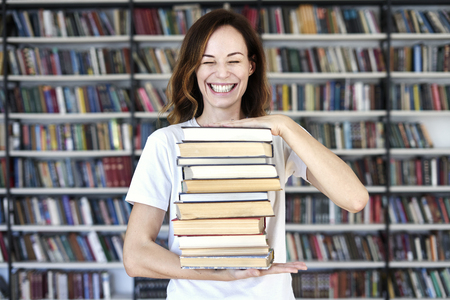 Woman model college student with books at library holds bunch of books, laughs looks smart, smiling to camera.の写真素材