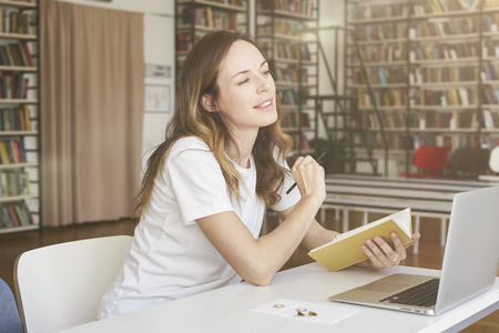 Young woman working on laptop at sunny co-working office or library, bookshelf behind. Businesswoman writing, looks smartの写真素材