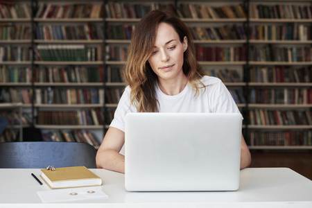 Woman entrepreneur working on laptop at co-working office or library, looks smart, bookshelves. Knowledge and self-development.の写真素材