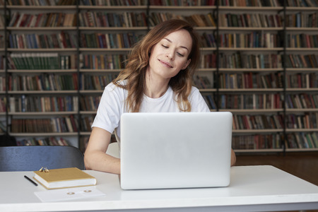 Woman entrepreneur with long hair working on laptop at co-working office or library, smiling dreaming, bookshelves. Knowledge and self-development.の写真素材