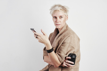Portrait of blond caucasian woman angry anxious emotion drinks takeaway coffee, using smartphone on white background.の写真素材