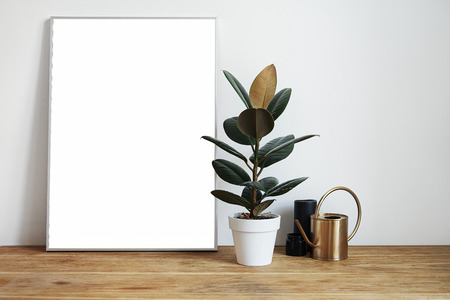 White room interior with green plants on rustic wooden table, modern personal laptop, poster in frame with space for layout.の写真素材