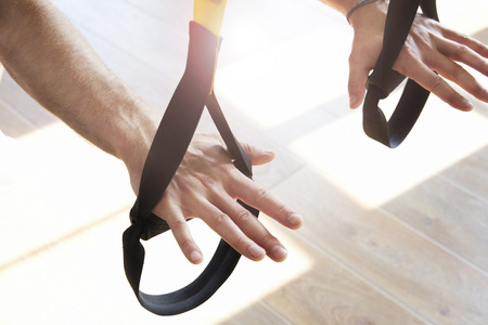 Close-up of man hands doing exercise alone with fitness belts in modern gym club, workout single sport, sun shining.の写真素材