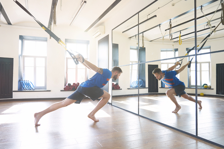 Handsome muscular man athlete doing exercise with fitness belts in modern sport club, sun shining through windows.の写真素材