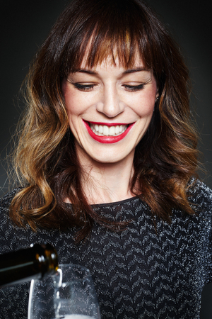 Woman wearing night party dress with a glass of champagne on dark background. Lady with long curly hair and perfect teeth celebrating and smiling. Pouring sparkling wineの写真素材