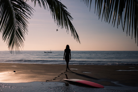 Female surfer with board on foreground walking along coastline on sandy beach over sunsetの写真素材