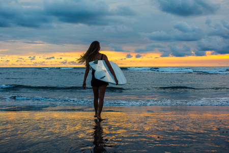 Female surfer with board in hands walking along coastline on sandy beach over sunsetの写真素材