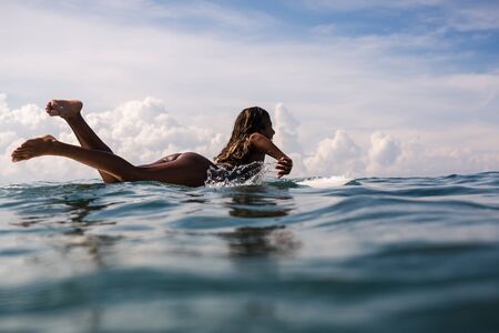 Beautiful young indonesian woman in bikini surfing wave in Bali on the background of blue sky, clouds and tropical beachの写真素材