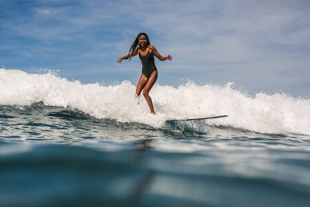 Beautiful young indonesian woman in bikini surfing wave in Bali on the background of blue sky, clouds and tropical beachの写真素材