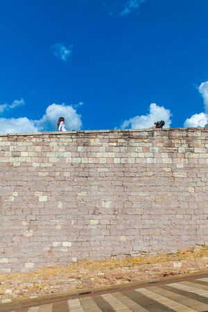 A boy punctuates a girl on the top of the wall in front of the Basilica of San Francesco in Assisi, Umbria, Italyの写真素材