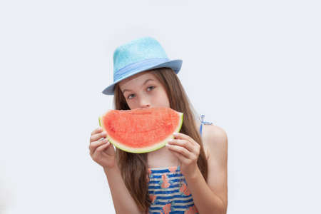 Young girl in a blue hat holding a slice of watermelon on a white background. Vegetarian girl eating watermelonの写真素材