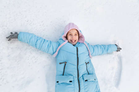 Portrait of a girl in winter in a pink hat and blue jacket. Girl lies on the snow. Place for text.の写真素材