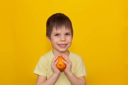 A cheerful child in a yellow T-shirt on a yellow background holds an orange in his hands. The concept of healthy food for children. copy spaceの写真素材