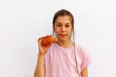 Playful cheerful red-haired girl with hair tied in ponytail holds two croissants in hands, looks at...