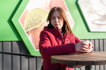 A woman stands near a round saltbox in a street cafe, holding a paper cup of coffee and looking...